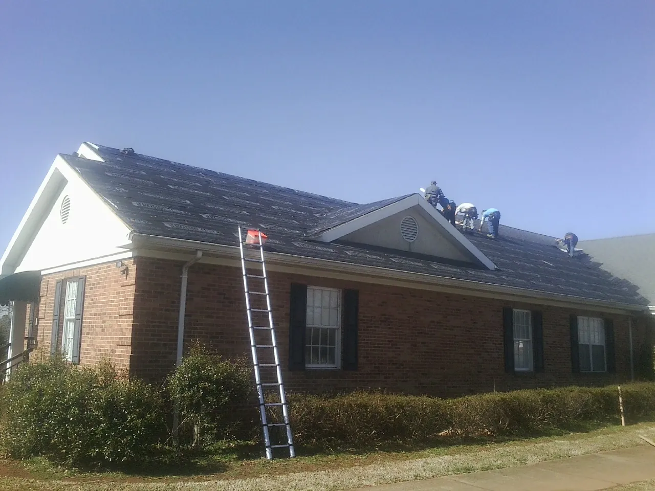 Crew tearing shingles off a brick ranch roof with a ladder set — Metal Roof Repair work by our crew in Minford