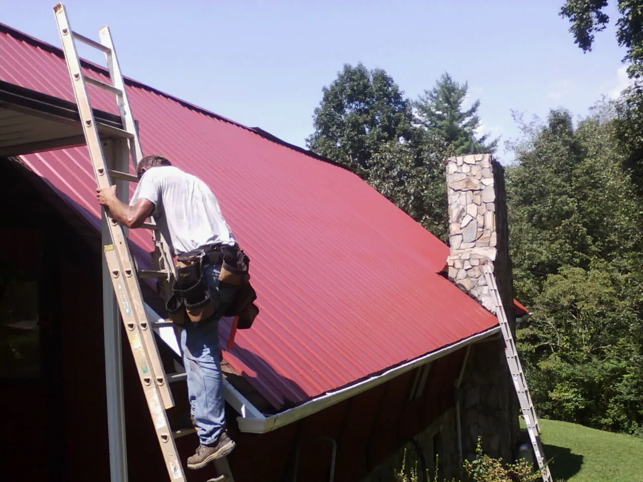 Roofer on a ladder beside a red metal roof and stone chimney — Soffit & Fascia Repair by Roofing Brothers in Minford
